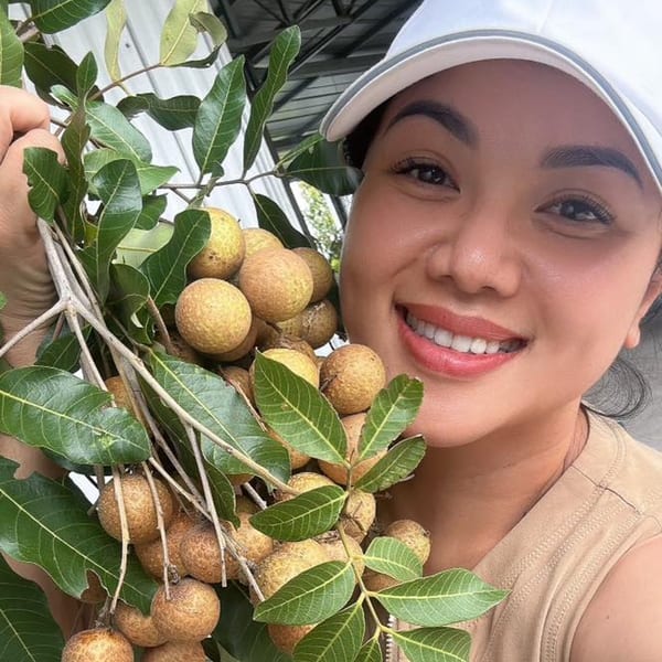 Smiling woman in a white cap holding a large bunch of fresh longan fruit with green leaves.