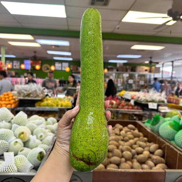 A hand holds a long green avocado in a brightly lit grocery store with produce and people.