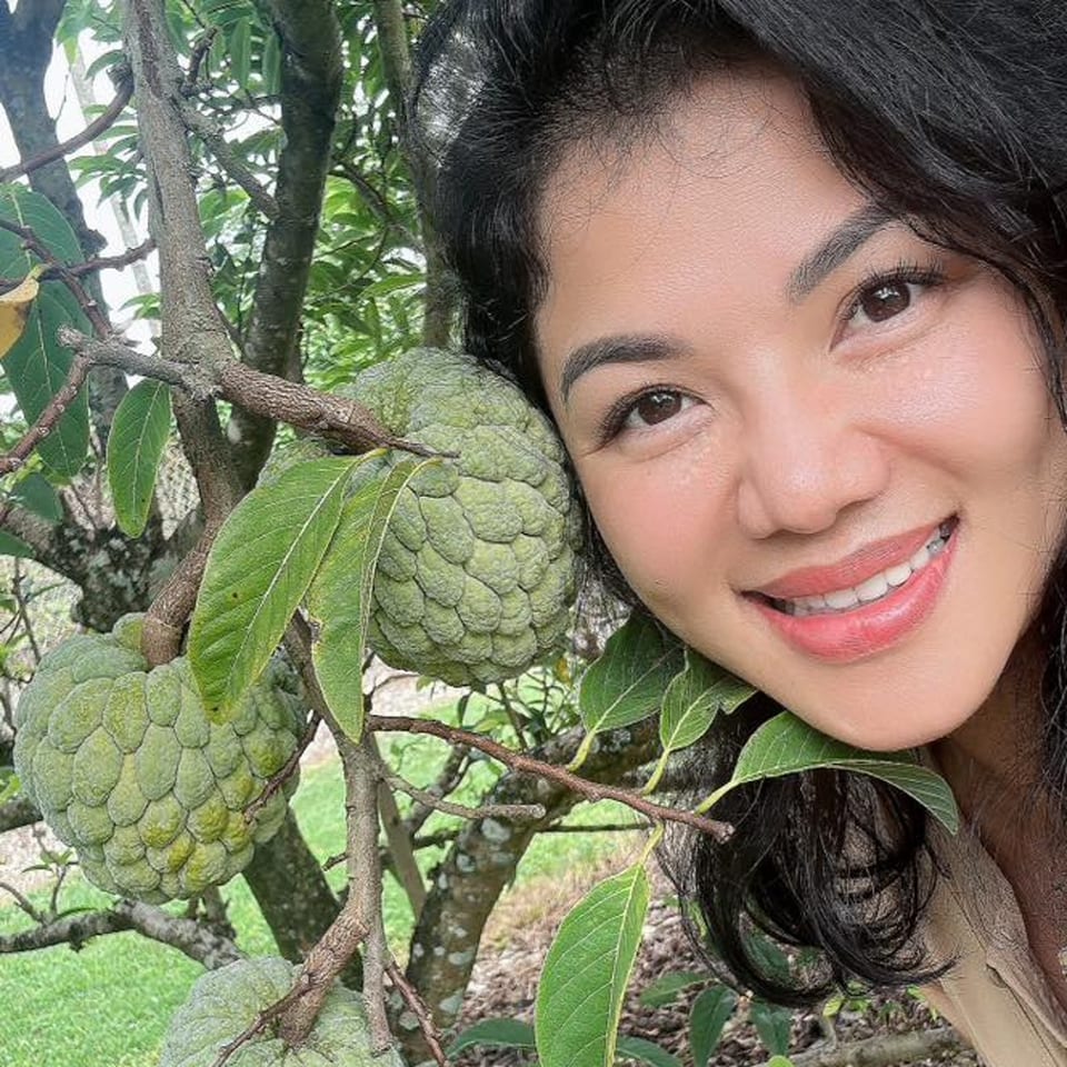A smiling woman with dark hair poses next to large, green soursop fruits on a tree.